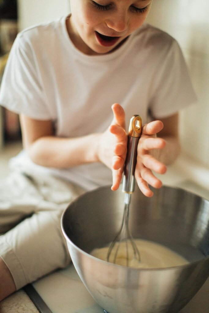 pexels-photo-3807115-3807115 A young boy happily using a whisk in a kitchen, expressing joy and fun while baking.